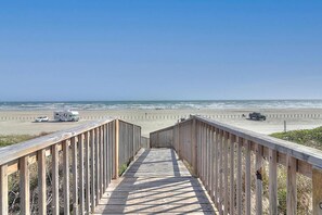 Ein hölzerner Steg führt zu einem Strand, an dessen Seite Autos geparkt sind.