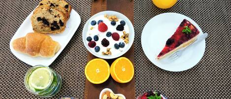 Ein Frühstückstisch mit Croissants, einem Stück Käsekuchen, Joghurt mit Beeren, einer herzförmigen Schüssel mit Mandeln, einer Schüssel mit bunten Beeren und einem Glas Orangensaft.