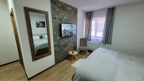 Habitación de hotel con cama, suelo de madera, pared de piedra, televisor montado, una pequeña mesa redonda con un teléfono y una ventana con cortinas.