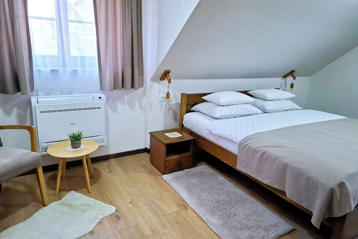 Une chambre à coucher avec un lit, une petite table sur laquelle est posée une plante, un radiateur et une fenêtre avec des rideaux.