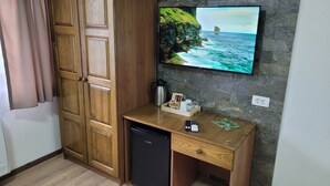 Habitación compacta con un armario de madera, un pequeño escritorio de madera con una cafetera, un control remoto y una revista. Un televisor montado en una pared de piedra muestra una escena costera.
