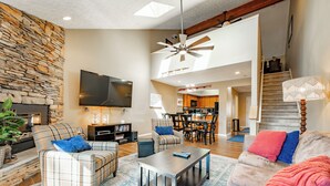 A modern living room with a stone wall, a flat-screen TV, a ceiling fan, and a staircase leading to a kitchen area. - Deep Creek Fun w/ Hot Tub, Dock & Fireplace (McHenry)