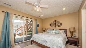 A bedroom with a bed, a nightstand, a ceiling fan, a window with curtains, and a view of a staircase leading outside. - Deep Creek Fun w/ Hot Tub, Dock & Fireplace (McHenry)