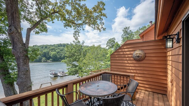 A wooden deck with a round table and chairs, overlooking a lake with boats and surrounded by trees. - Deep Creek Fun w/ Hot Tub, Dock & Fireplace (McHenry)