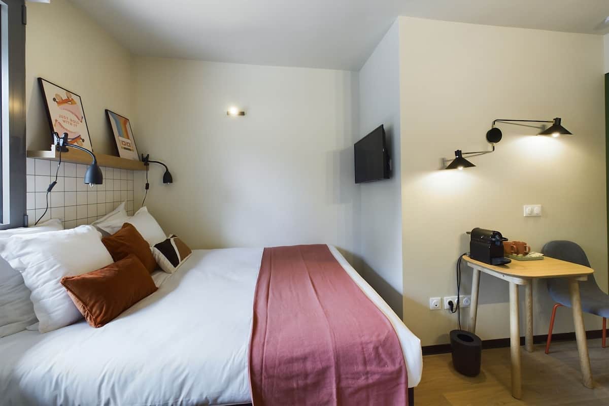 A hotel room with a bed, a TV mounted on the wall, a desk with a chair, and a small table with a coffee maker.