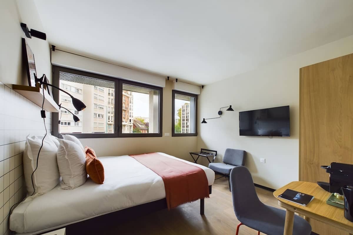A modern hotel room with a bed, a desk, a chair, a TV, and large windows.
