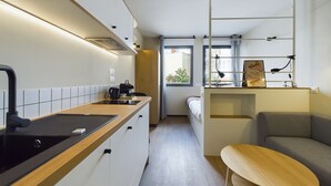 A compact, modern kitchenette with a built-in stove, a small sink, and a countertop. There is a bed with a gray headboard and a wooden bench in the adjacent room. - Hife Paris Gentilly (Gentilly)