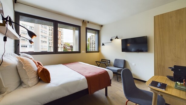 A modern hotel room with a bed, desk, chairs, a TV, and large windows. - Hife Paris Gentilly (Gentilly)