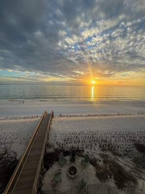 A wooden boardwalk leading to a sandy beach with a fire pit and chairs.
