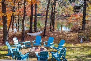 Een vuurkorf omringd door blauwe Adirondack-stoelen in een bebost gebied met een vijver.