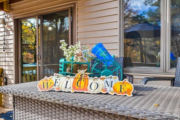 Een rieten tafeltje met een 'WELCOME' bord, versierd met pompoenen en bloemen, voor een huis met ramen.