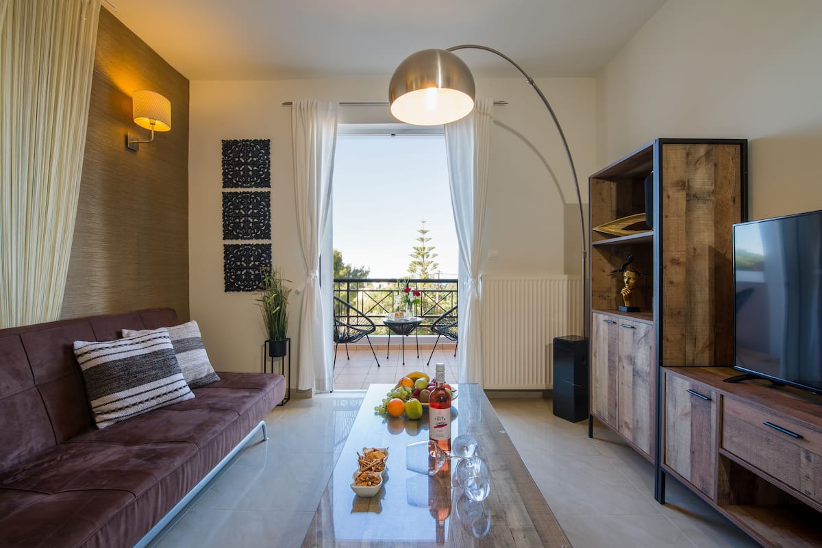 Un salon moderne avec un canapé, une table basse sur laquelle sont disposés des fruits et des en-cas, un téléviseur et un balcon offrant une vue agréable.
