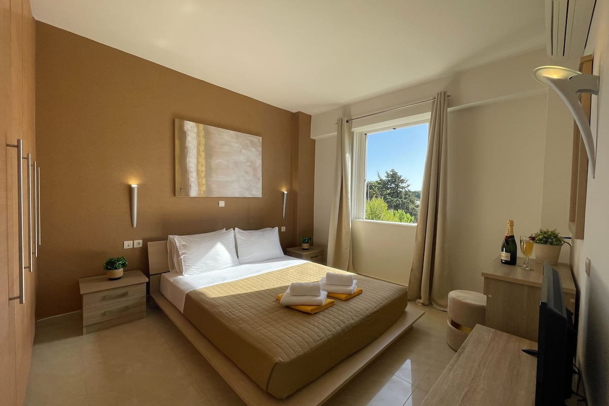 Une chambre moderne avec un grand lit, des tables de chevet, une fenêtre avec des rideaux et un petit bureau sur lequel se trouvent une bouteille et un verre.