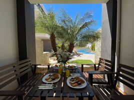 Un área de comedor al aire libre con dos mesas, cada una dispuesta con platos de comida, cubiertos y vasos. Se observan palmeras y una piscina al fondo.