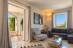 A modern living room with a sofa, armchair, glass coffee table, and a view of greenery outside. - Artdeco Luxury Suites (Chania)