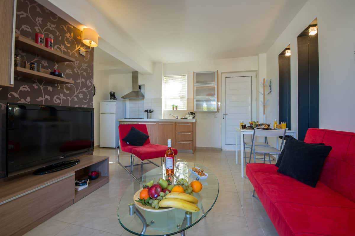 Un salon moderne avec une table basse en verre, un canapé rouge et un téléviseur. On aperçoit une cuisine à l’arrière-plan, avec un coin repas.