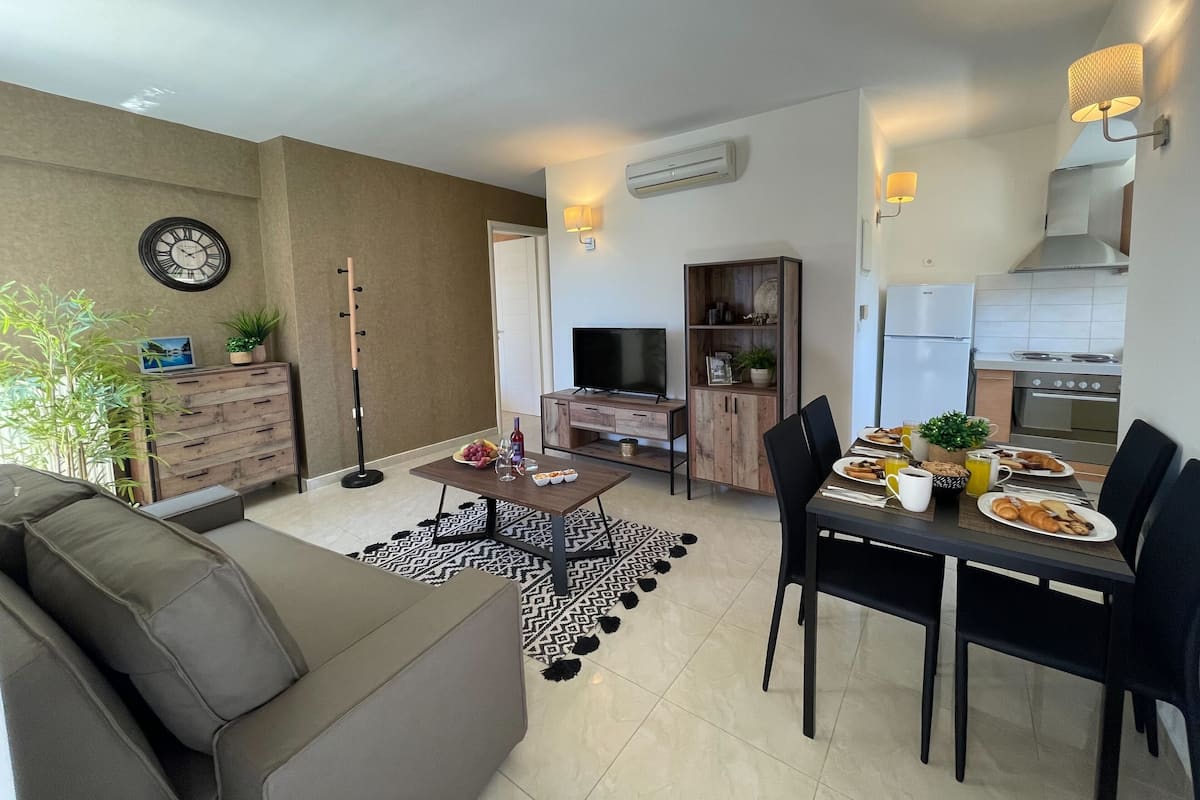Un salon moderne comprenant un canapé, une table à manger et des chaises. On y trouve également un téléviseur, un meuble en bois et un coin cuisine avec une cuisinière et un réfrigérateur.