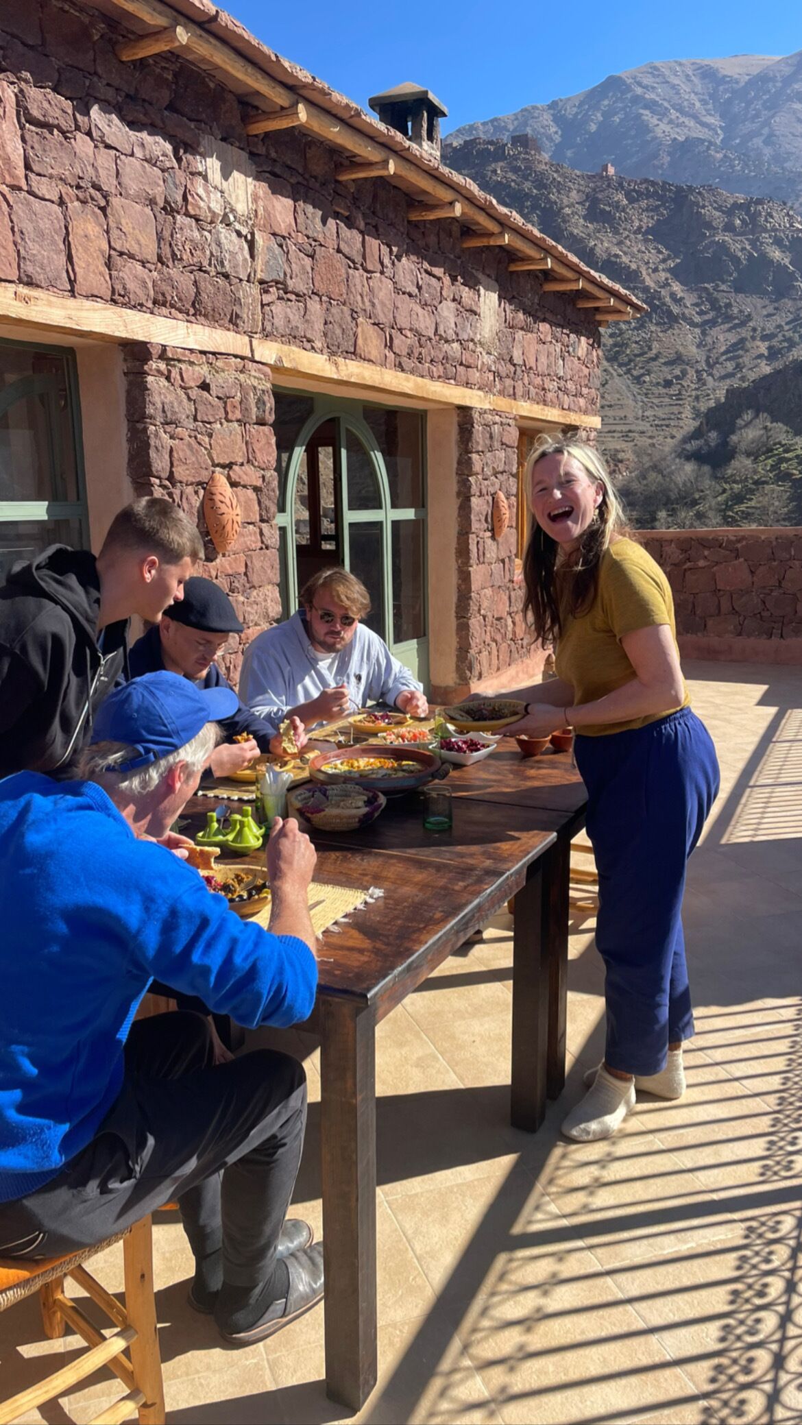 A group of people dining outdoors at a rustic stone building with a mountain backdrop.