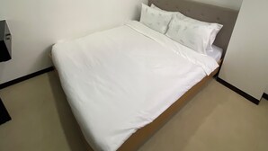 Un lit simple avec une literie blanche, dans une pièce aux murs unis et dotée d’une tête de lit en bois.