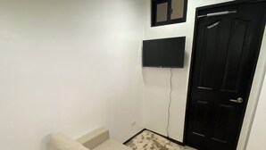 Une pièce avec un canapé beige, un téléviseur à écran plat fixé au mur, une porte noire et un tapis à motifs sur le sol.