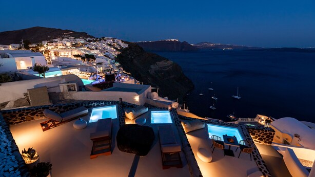 A seaside resort with a pool area, lounge chairs, and a view of the ocean and distant hills. - Twenty One Suites (Santorini)