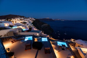 A seaside resort with a pool area, lounge chairs, and a view of the ocean and distant hills. - Twenty One Suites (Santorini)