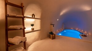 A spa room with a wooden shelf holding towels, a small plant, and a basket with bath products. A blue-lit pool is visible in the background. - Twenty One Suites (Santorini)