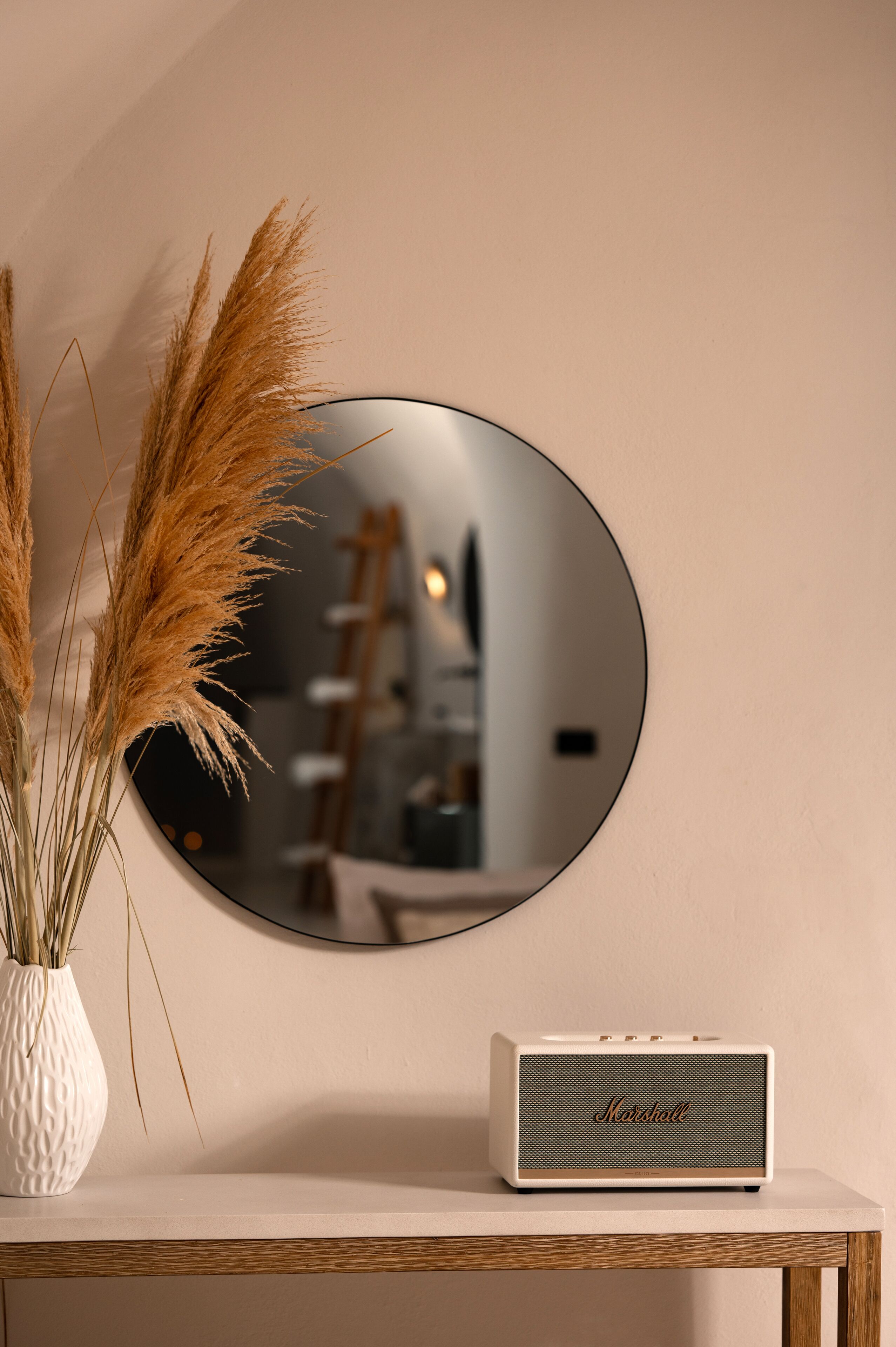 A white Marshall speaker on a wooden shelf, a white vase with pampas grass, and a round mirror on a beige wall.