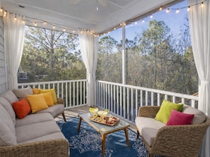 Alpendre coberto com móveis de vime, mesa de jantar com um prato de frutas e vista para árvores através de grandes janelas.
