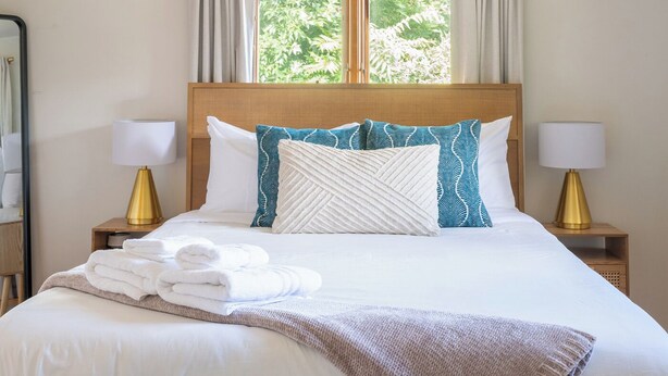 A neatly made bed with white linens, a brown throw blanket, and decorative pillows. There are two bedside lamps with gold bases and white shades. A window with curtains allows natural light into the room. - Cozy Westminster Retreat ~ Hot Tub ~ Near Denver ~ Spacious Yard (Westminster)