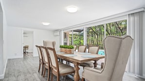 A dining area with a wooden table and chairs, a small plant centerpiece, and a view of a pool through large windows. - Naples Dream Escape 4BR Home, heated pool, 10min from Beach (Naples)