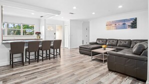 A modern living room with a sectional sofa, a dining area with bar stools, and a large window with a view of palm trees. - Naples Dream Escape 4BR Home, heated pool, 10min from Beach (Naples)