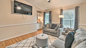 A living room with a flat-screen TV, a ceiling fan, a sofa, a coffee table, and a window with curtains. - Lillington Home ~ 1 Mi to Cape Fear River! (Lillington)