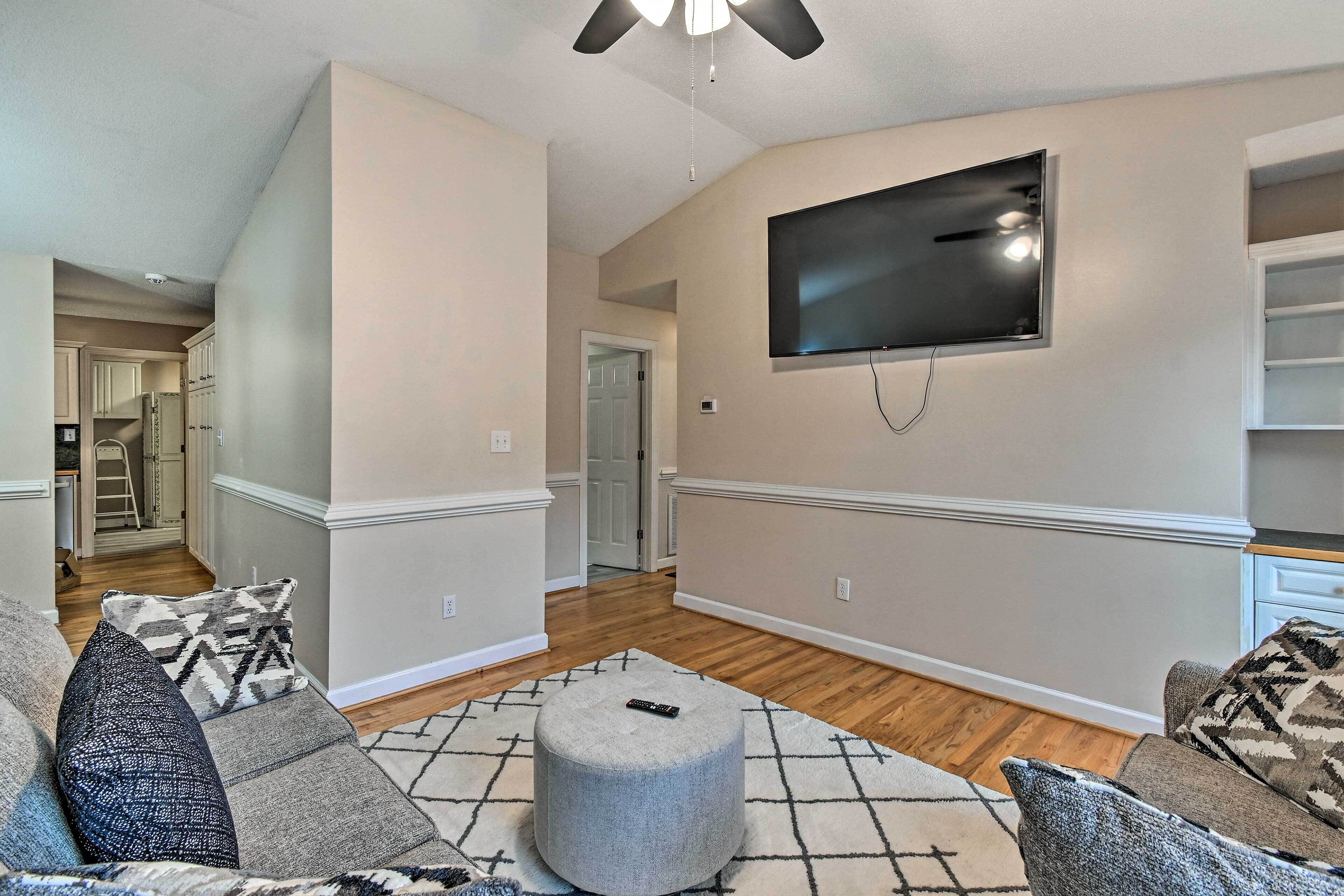 A living room with a large flat-screen TV mounted on the wall, a grey ottoman, and a patterned rug.