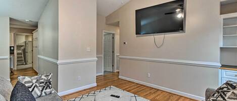 A living room with a large flat-screen TV mounted on the wall, a grey ottoman, and a patterned rug.