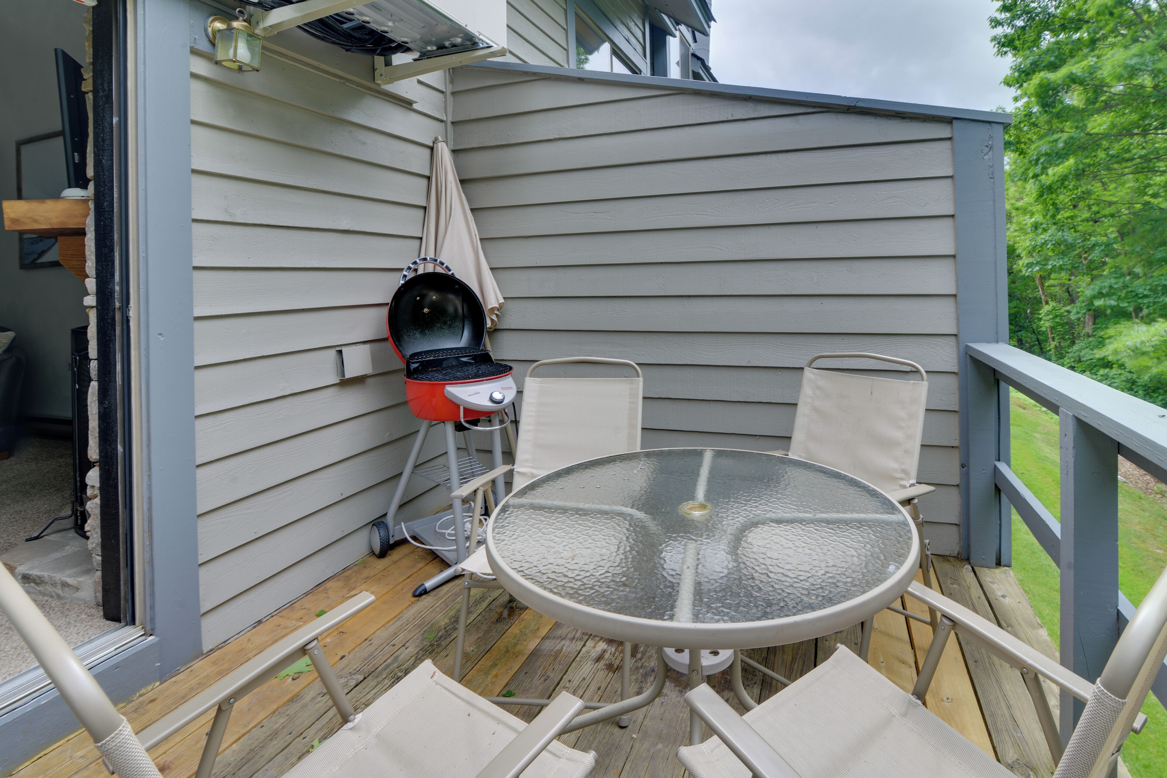 A covered outdoor patio with a glass table and metal chairs, a barbecue grill, and a wooden deck.