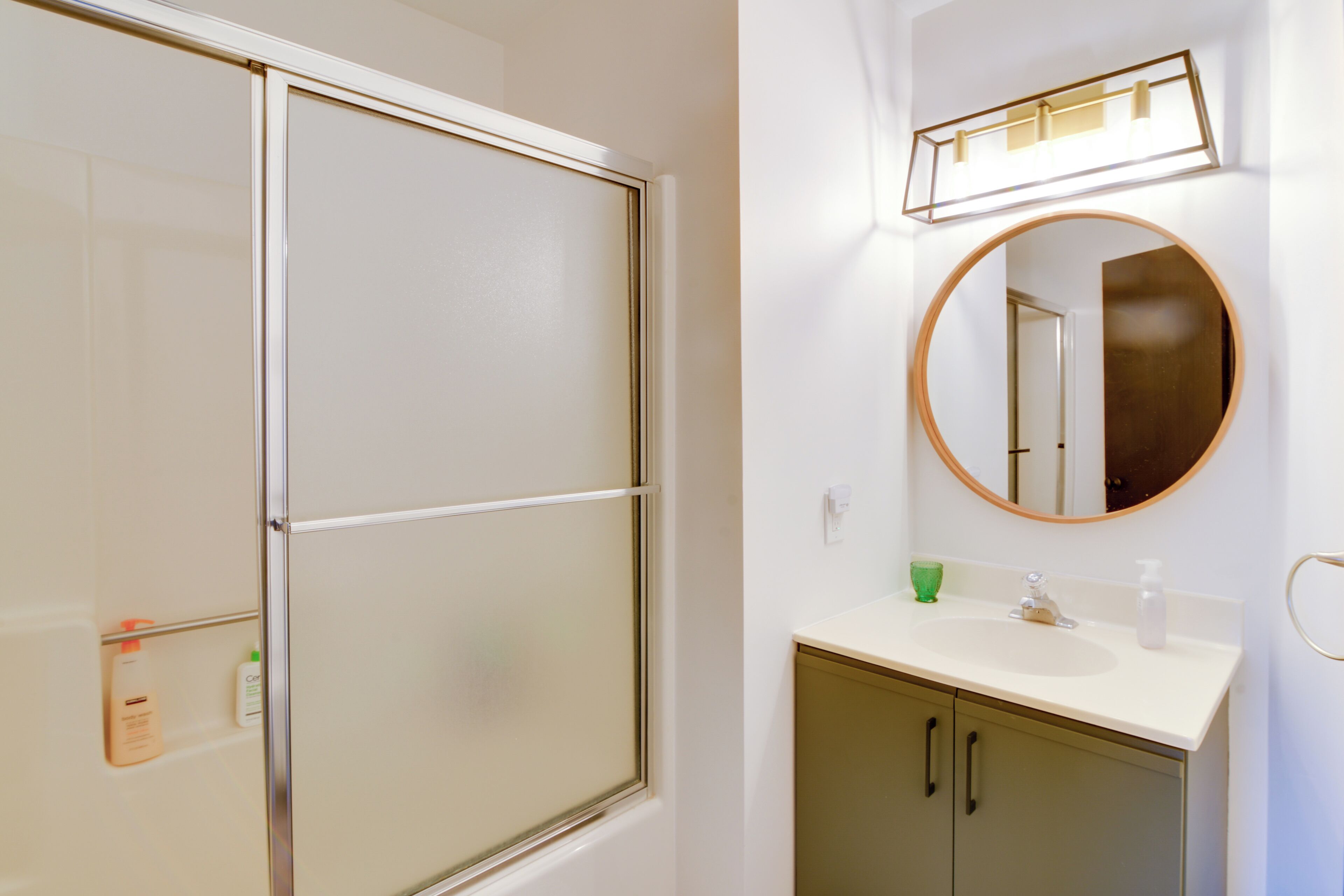 Een badkamer met een deur van matglas voor de douche, een ronde spiegel, een wit wastafelbakje en een groene beker op het aanrecht.