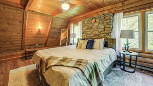 A cozy wooden cabin bedroom with a stone wall, a bed with multiple pillows, a ceiling fan, and a window with curtains. - Hot Tub, Fire Pit: Waynesville Getaway Near Hiking (Canton)