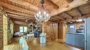 A log cabin interior with a wooden dining table, white chairs, a chandelier, a kitchen area, and a cozy fireplace. - Hot Tub, Fire Pit: Waynesville Getaway Near Hiking (Canton)