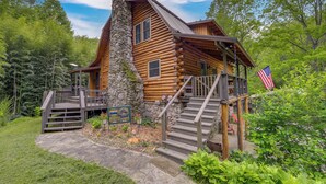 A log cabin with a stone chimney, wooden deck, and a flag. - Hot Tub, Fire Pit: Waynesville Getaway Near Hiking (Canton)
