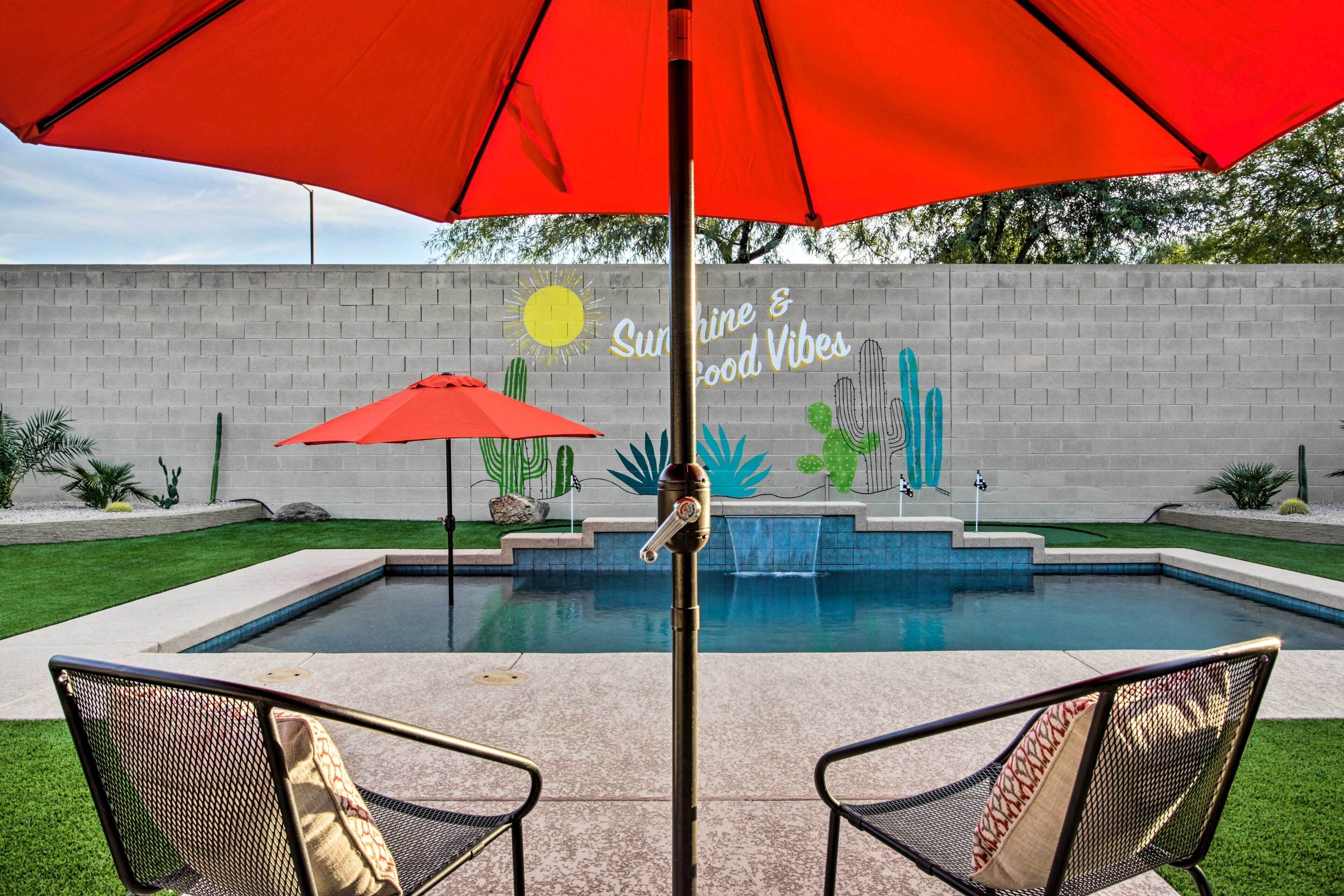 A pool area with two chairs and a red umbrella, a wall with a mural, and a water feature.