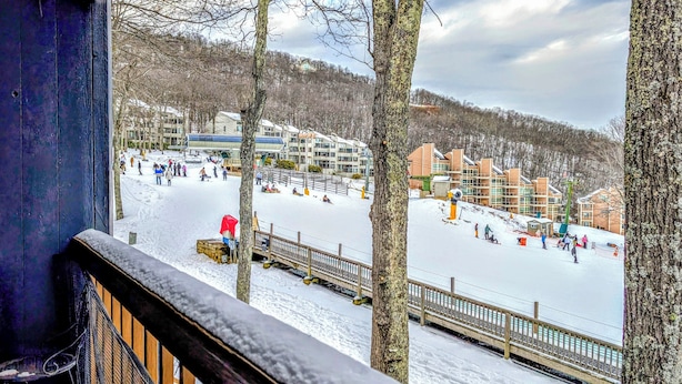 A snow-covered outdoor area with a wooden deck and railings, surrounded by buildings and trees.