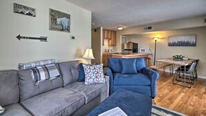 A living room with a grey sofa, blue armchair, and ottoman, a dining area with a wooden table and chairs, and a kitchen in the background.
