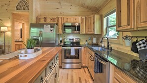 Cocina de cabaña de madera con refrigerador, horno y microondas de acero inoxidable, armarios de madera, encimera de granito y ventana con vistas a la vegetación.