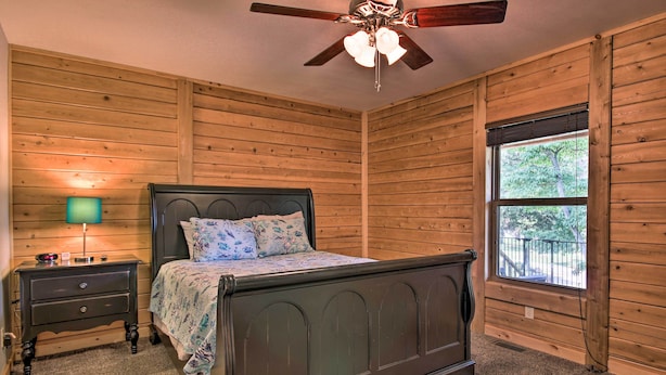 A bedroom with a wooden paneled wall, a bed with a headboard, a nightstand with a lamp, and a window with a view of trees. - 'rukarasa Cabin' w/ Yard & Dock at Grand Lake (Eucha)