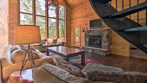 A cozy cabin interior with a stone fireplace, a large window, and a spiral staircase. - 'rukarasa Cabin' w/ Yard & Dock at Grand Lake (Eucha)