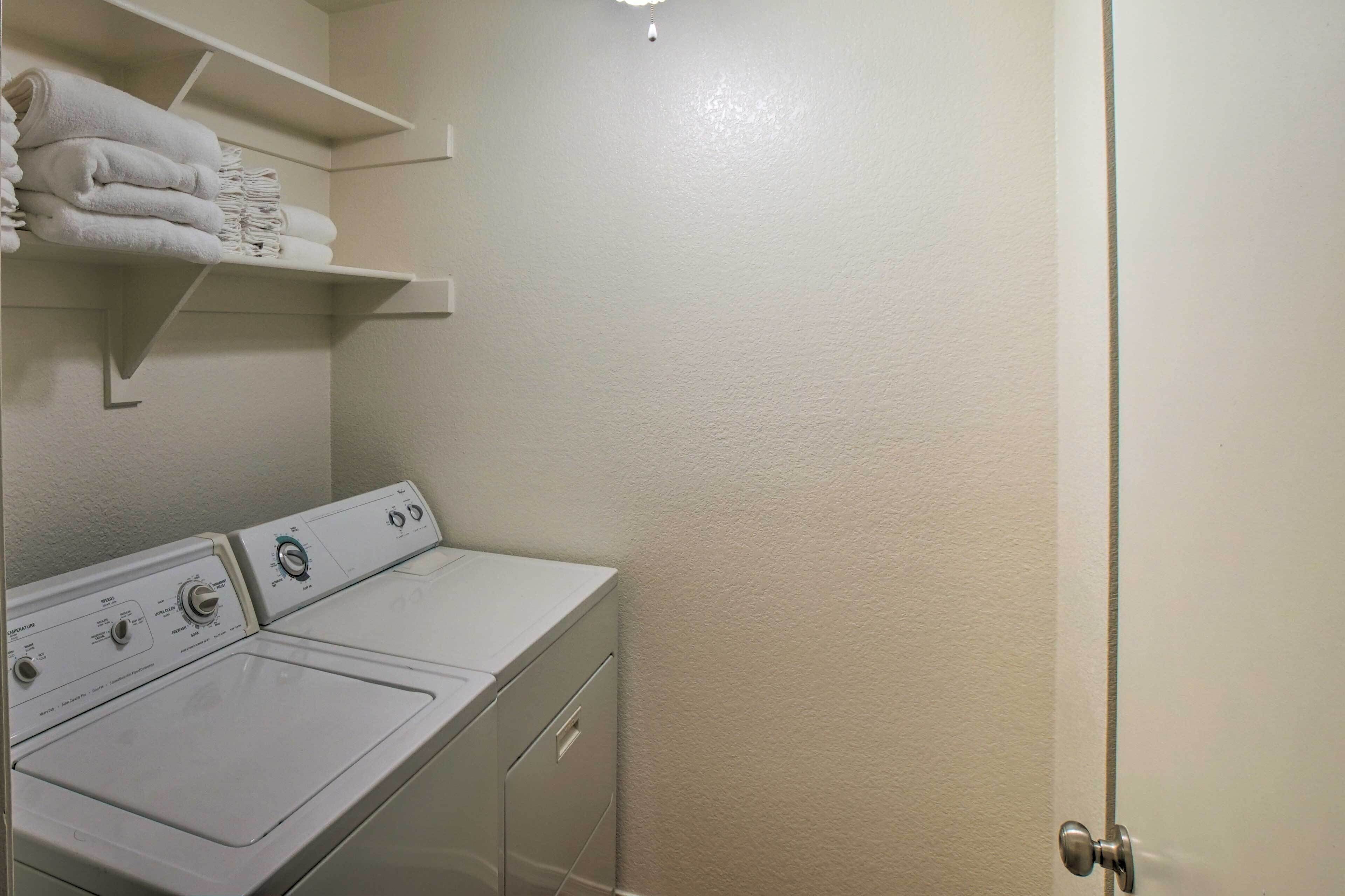 A laundry room with a front-loading washer and dryer, a shelf with folded towels, and a door to the right.