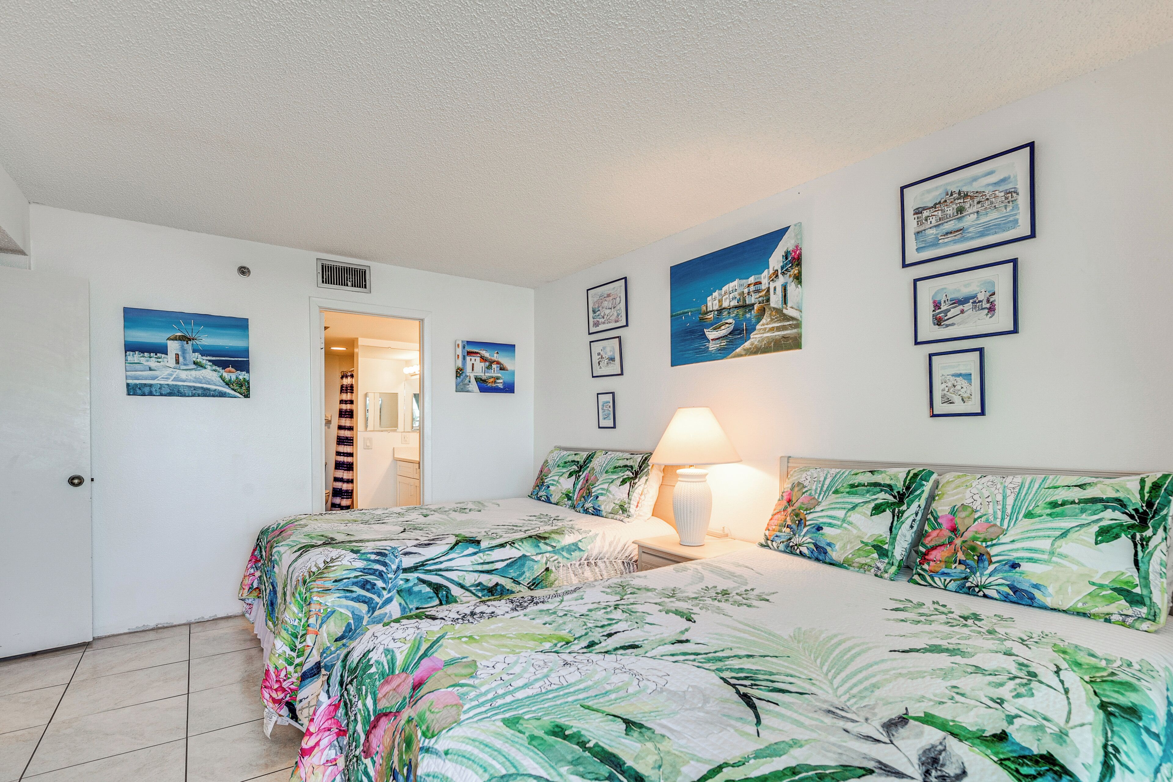 A bedroom with a bed, a bedside table, and a lamp. There are wall art pieces and a door leading to another room.