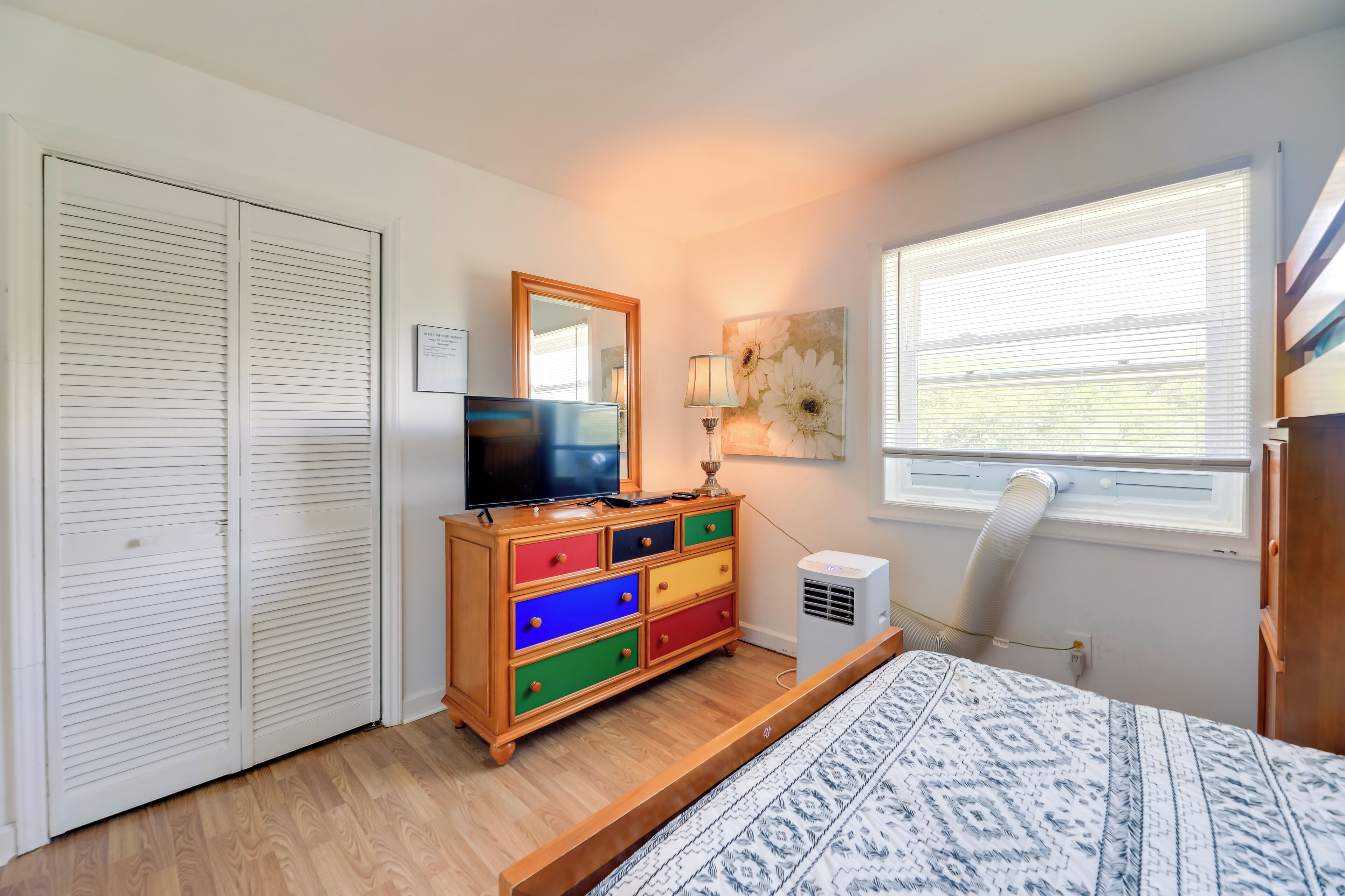 A bedroom with a bed, a dresser with colorful drawers, a television, a mirror, a window with blinds, and a wall-mounted air conditioner.