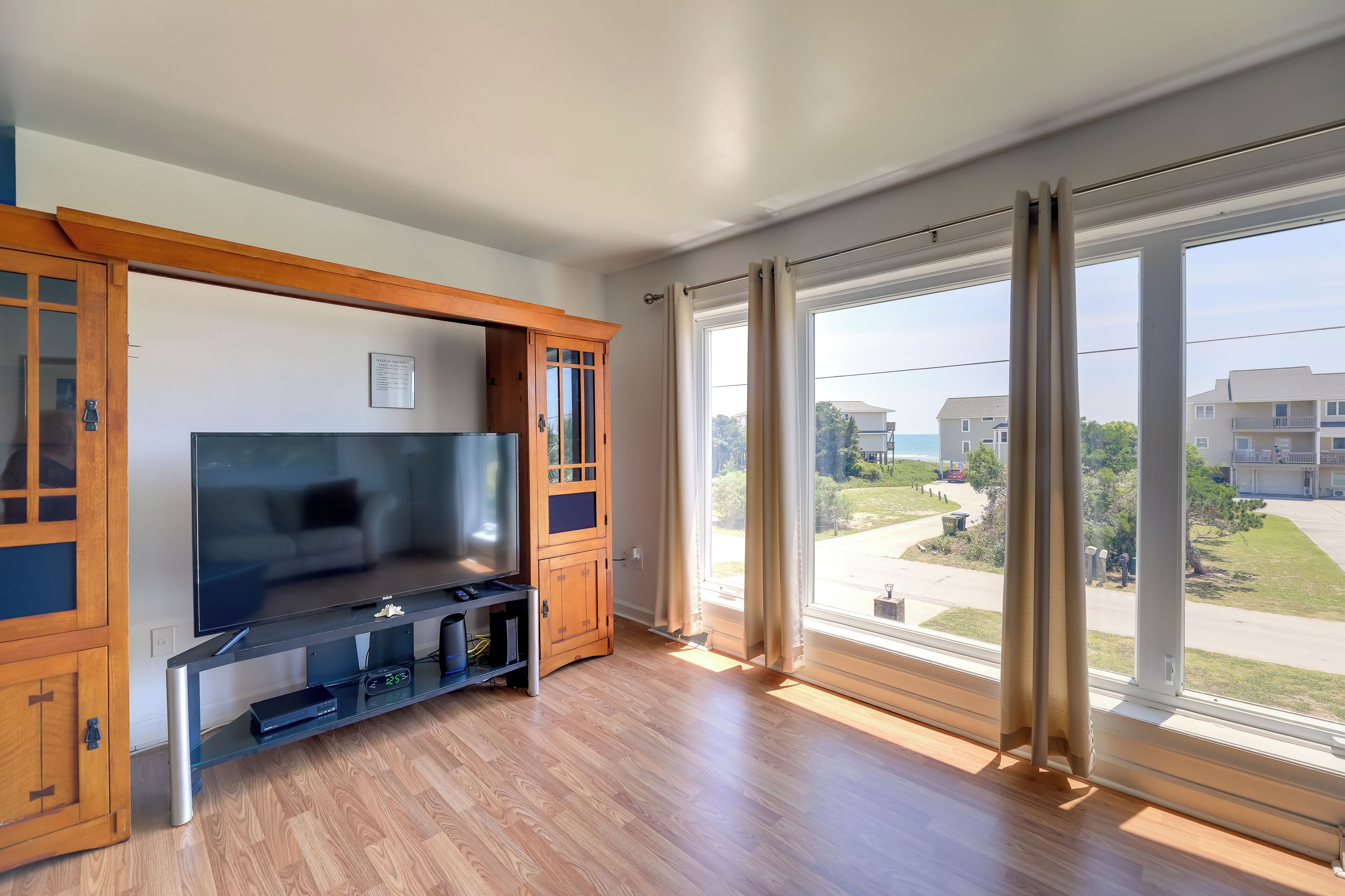 A modern living room with a large flat-screen TV, wooden cabinets, and a large window with a view of a residential area.